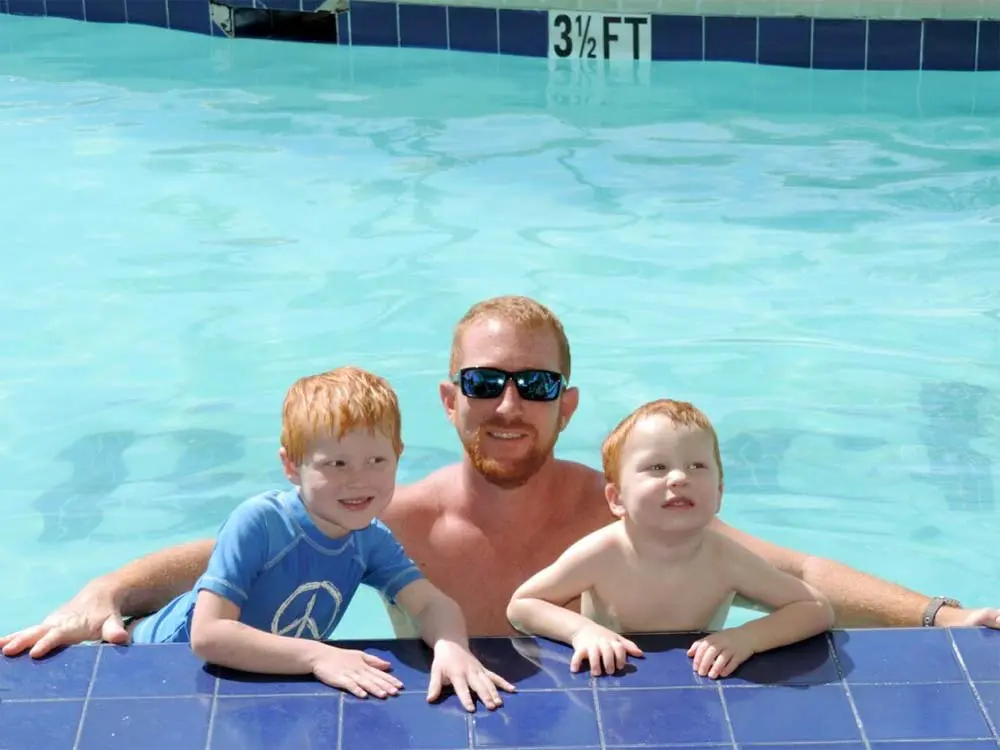 A man and two young children posing together in a swimming pool with a depth marker indicating 3 1/2 feet.