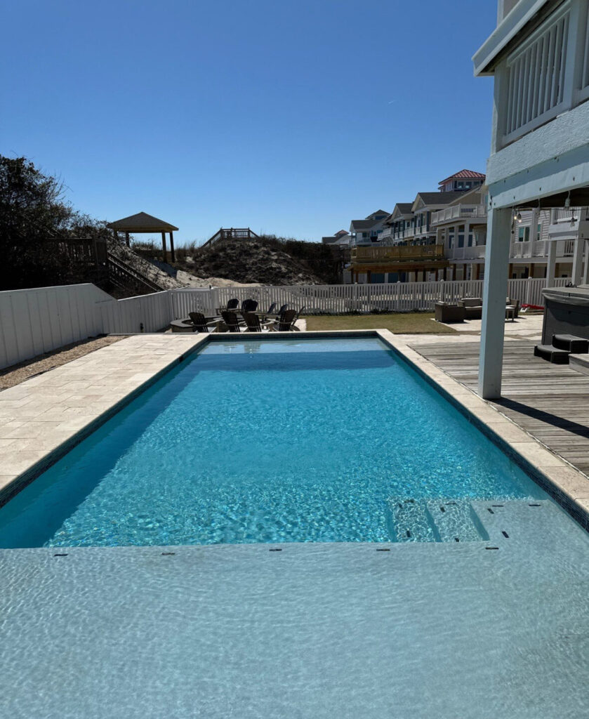 Rectangular outdoor swimming pool with clear water, surrounded by lounge chairs, deck, and nearby beach houses under a clear sky.