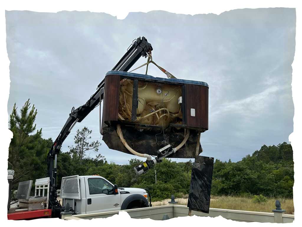 A crane attached to a truck lifts an old, damaged hot tub outdoors with trees in the background.