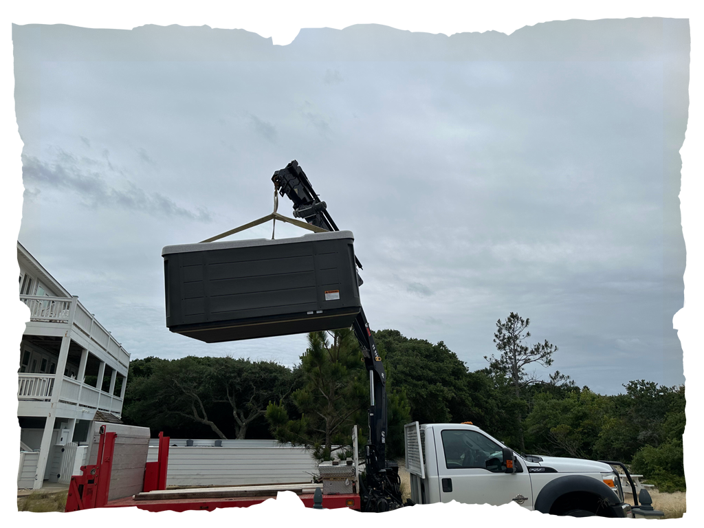 A crane truck lifts a large rectangular container near a white building and trees under a cloudy sky.