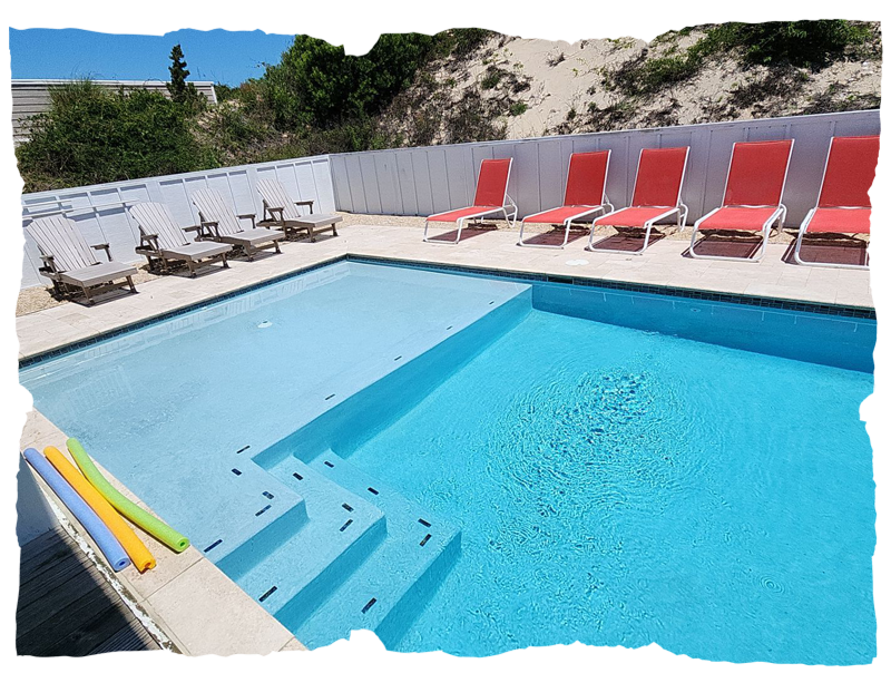 Outdoor swimming pool with clear water, red lounge chairs, wooden loungers, and pool noodles on the deck.