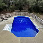 A fenced backyard pool with blue water, surrounded by lounge chairs and trees in the background.