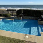 Rectangular outdoor pool with attached hot tub, surrounded by a patio, fence, and view of the ocean in the background.