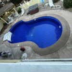 A small backyard swimming pool with blue tiles, surrounded by patio furniture and a hot tub on a concrete deck.