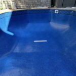An empty in-ground swimming pool with a blue liner, located in a backyard near a house and hot tub.