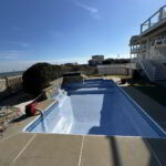 An empty in-ground swimming pool under repair sits in a fenced backyard with houses and blue sky in the background.