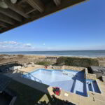 View of an empty backyard swimming pool near a beach, with clear skies and ocean in the background.