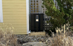 Freestanding black outdoor refrigerator placed between buildings on a wooden platform surrounded by rocks and vegetation.