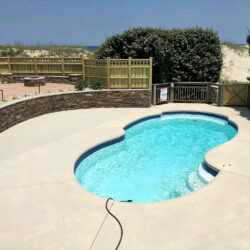 A kidney-shaped swimming pool with clear blue water, surrounded by a concrete deck and a wooden fence, with sand dunes and vegetation visible in the background.