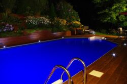 Illuminated backyard pool at night with surrounding garden and wooden deck.