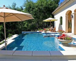 A sunlit backyard with a swimming pool, patio umbrellas, and a well-manicured garden beside a house.