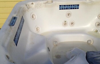 Empty, worn-out hot tub with visible grime and discoloration against a yellow wall.