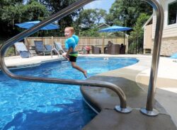 A child jumps into a swimming pool on a sunny day, framed by a stainless steel pool ladder.